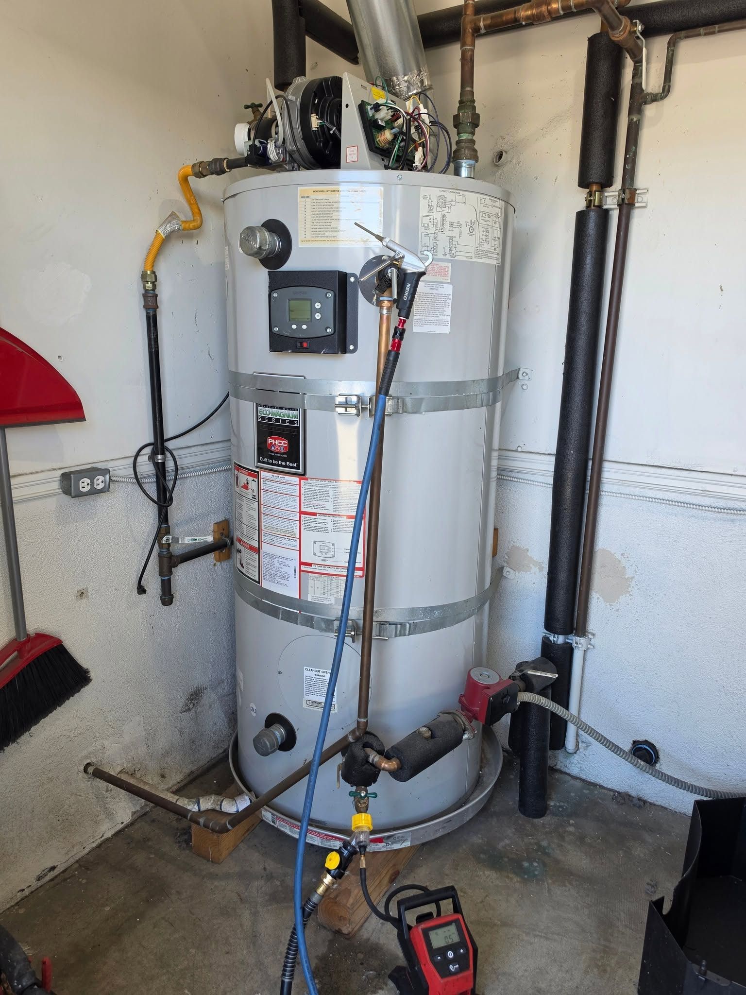 Water heater in a garage with visible pipes, valves, and a red floor pump nearby.