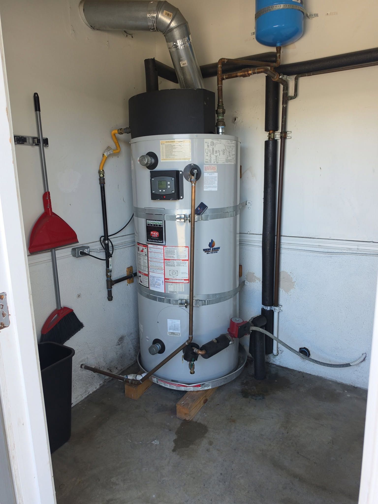 White water heater in a utility room with pipes, tank, and cleaning tools nearby