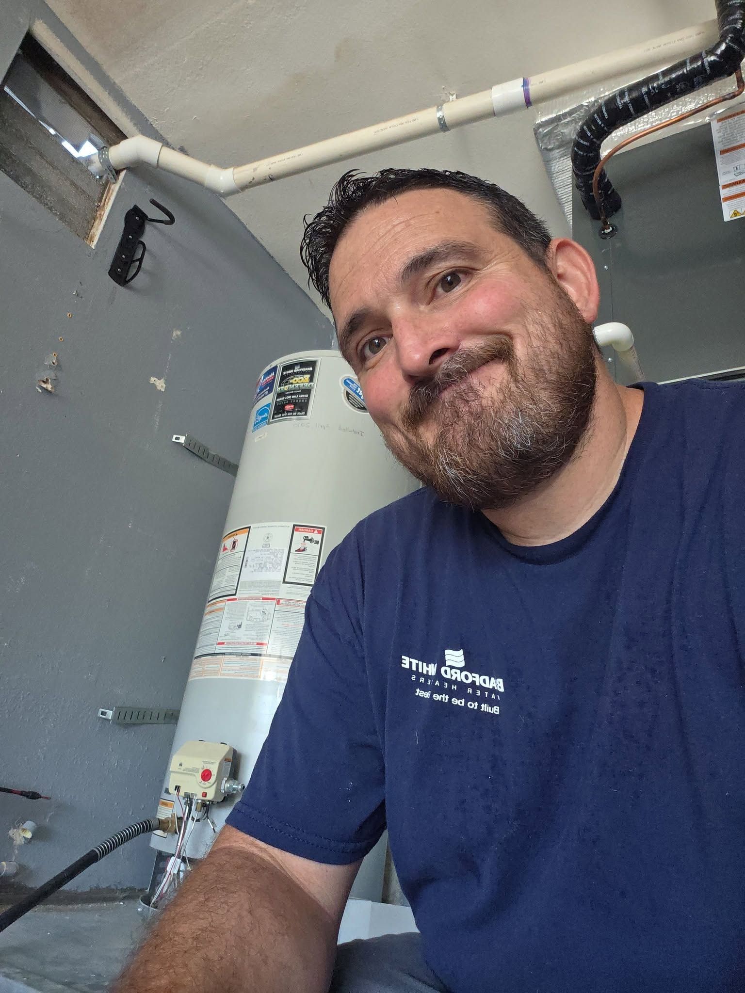 Man in a blue shirt smiling in an industrial room with pipes and a water heater behind him