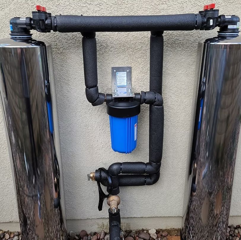 Blue water filtration system with black pipes and two stainless steel tanks mounted on a wall.