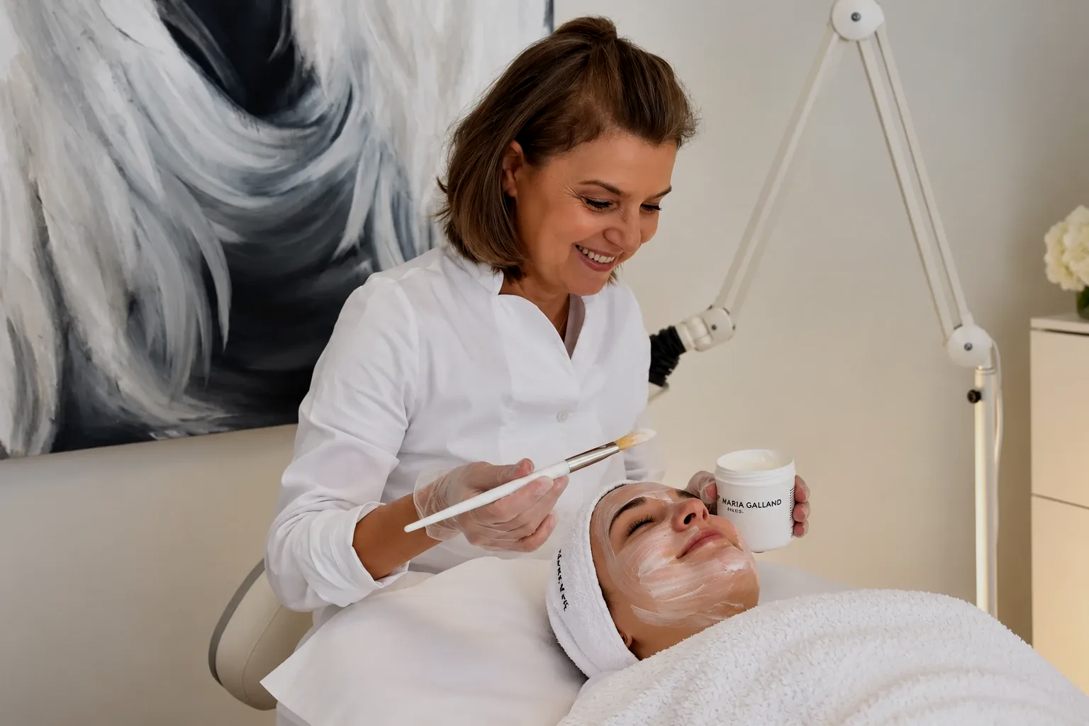 Eine Frau führt in einem hellen Behandlungsraum auf einer Spa-Liege eine Gesichtsbehandlung an einer Kundin durch.