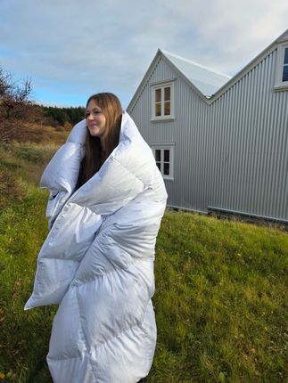Person wrapped in a large duvet standing outdoors beside a corrugated metal house.