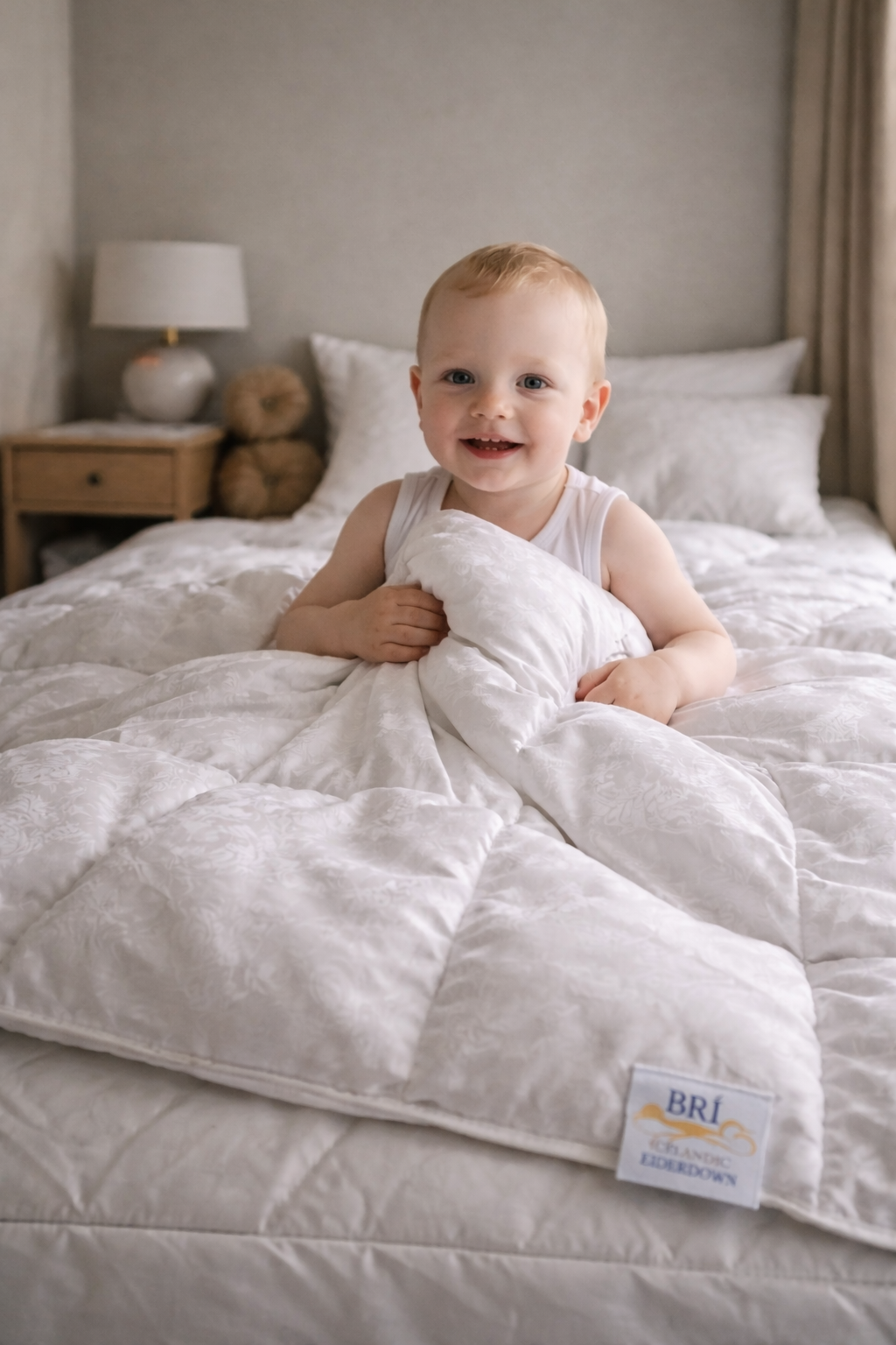 Smiling baby sitting on a white bed, holding a fluffy eiderdown duvet in a softly lit bedroom