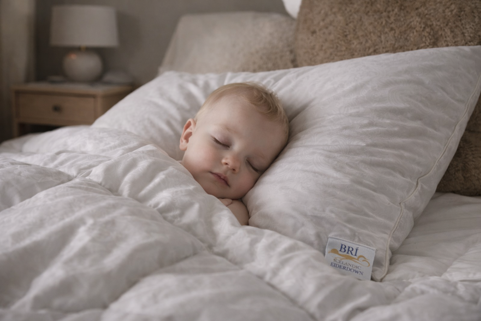 Smiling baby sitting on a white bed, holding a fluffy eiderdown duvet in a softly lit bedroom