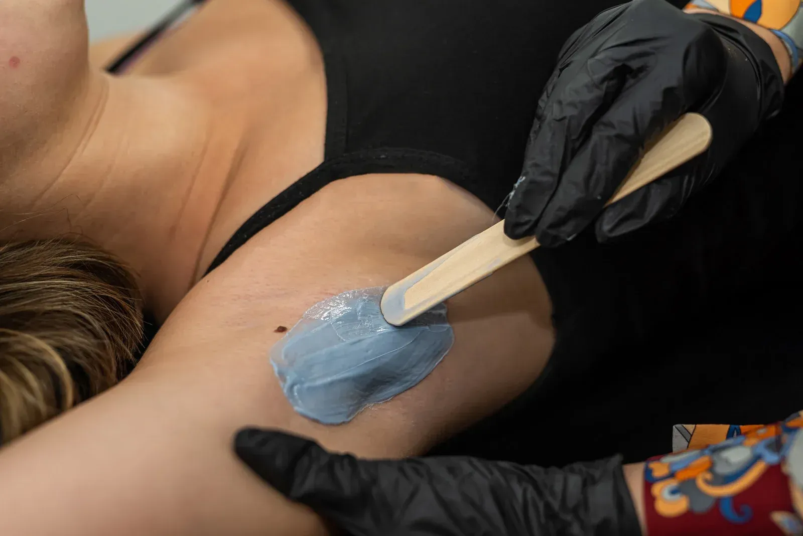A woman is getting her armpit waxed by a person wearing black gloves.