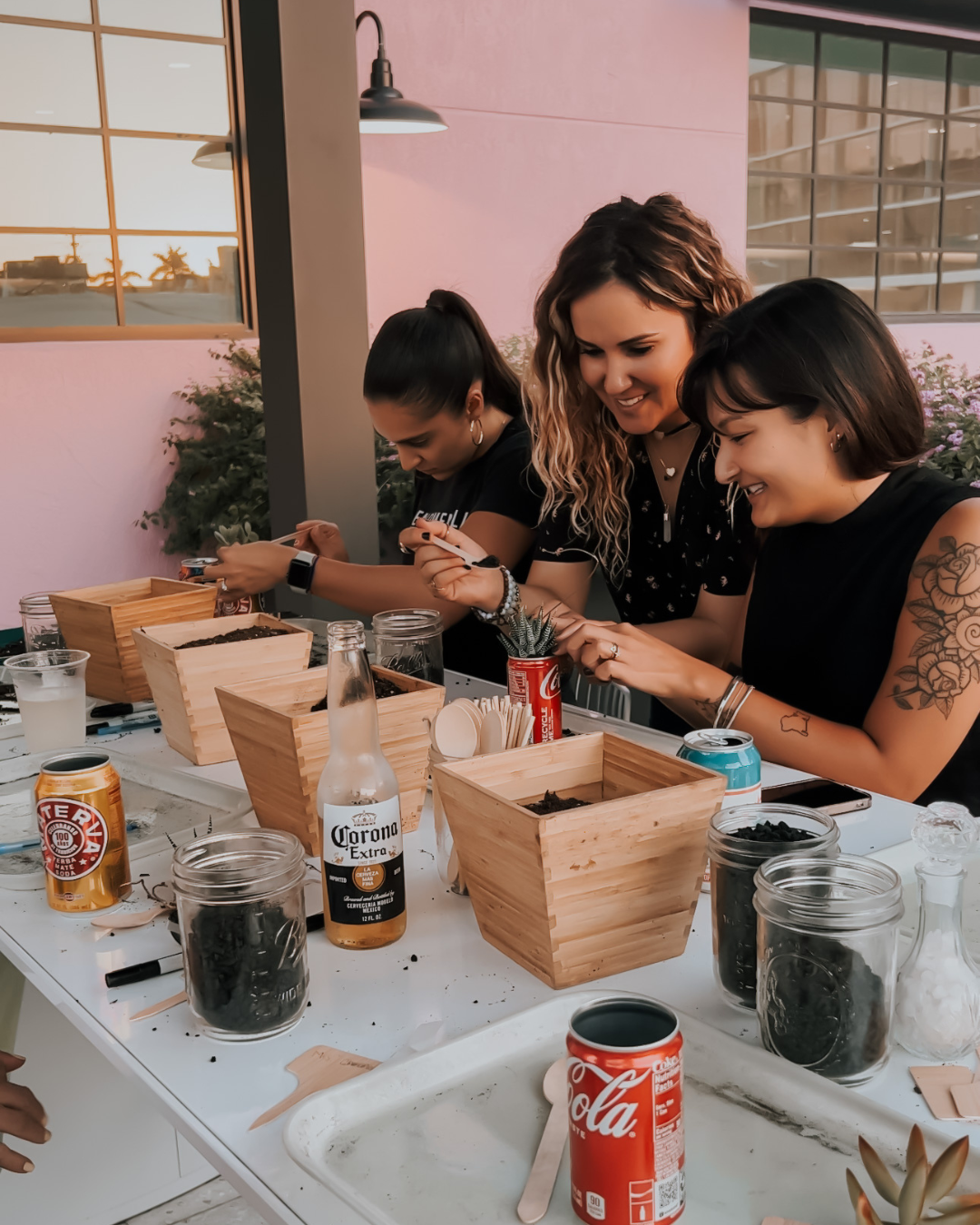Three women seated at a table, crafting with supplies and jars in a bright indoor workshop.