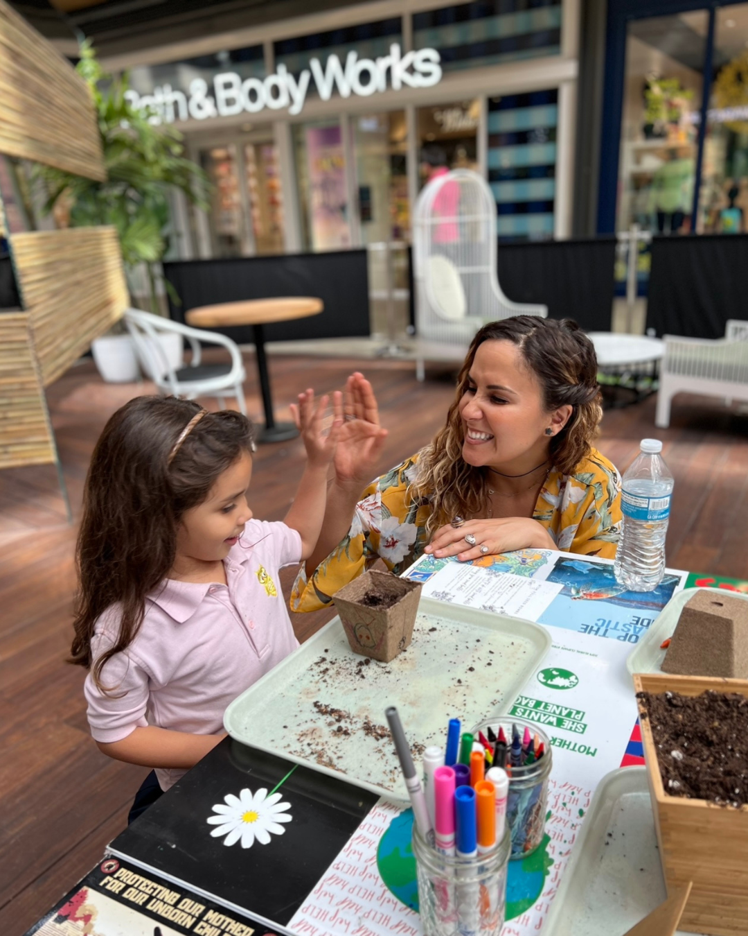 Two people high-five at a craft table with potted seedlings and markers in a store courtyard.