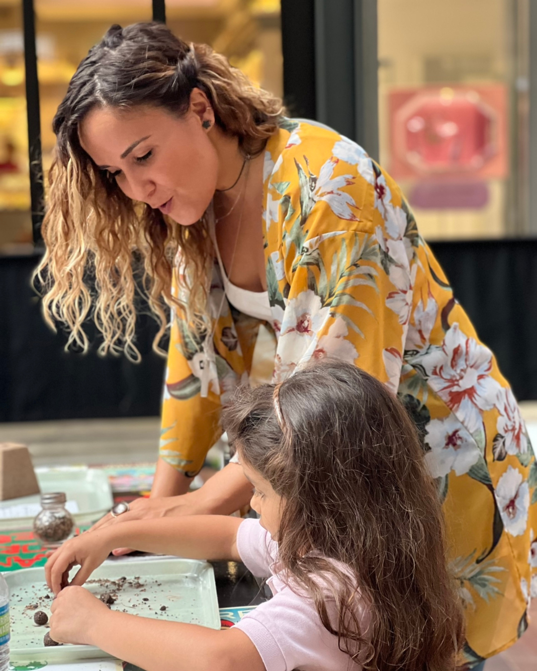 Woman and child painting together at a table, with the woman leaning over to help.