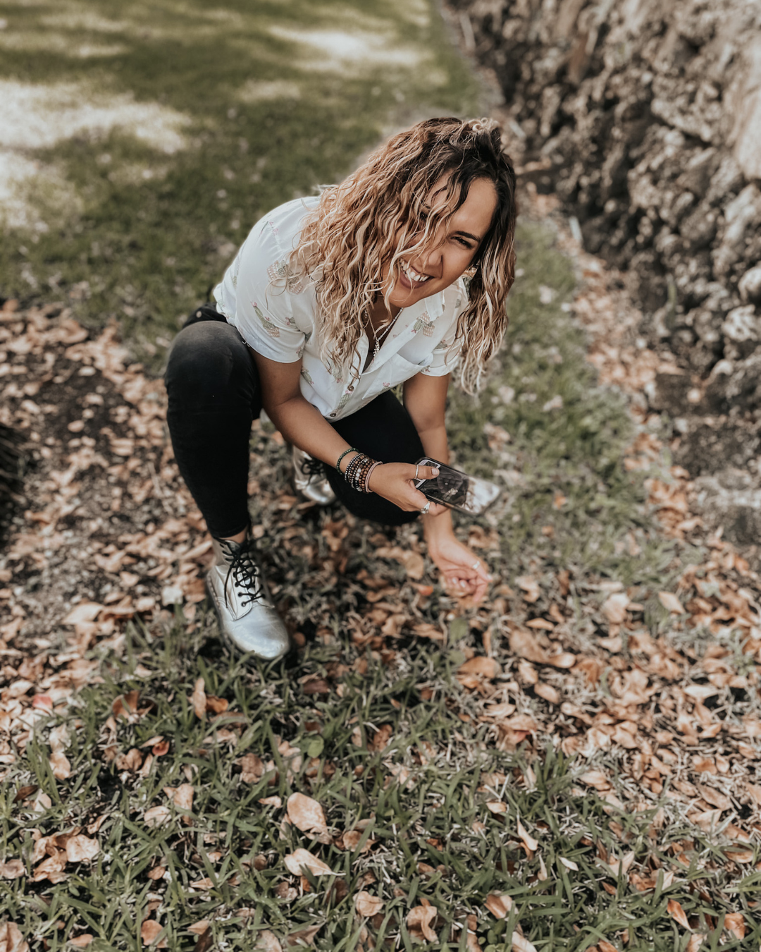 Woman crouching by a stone wall, collecting leaves on a grassy path outdoors.