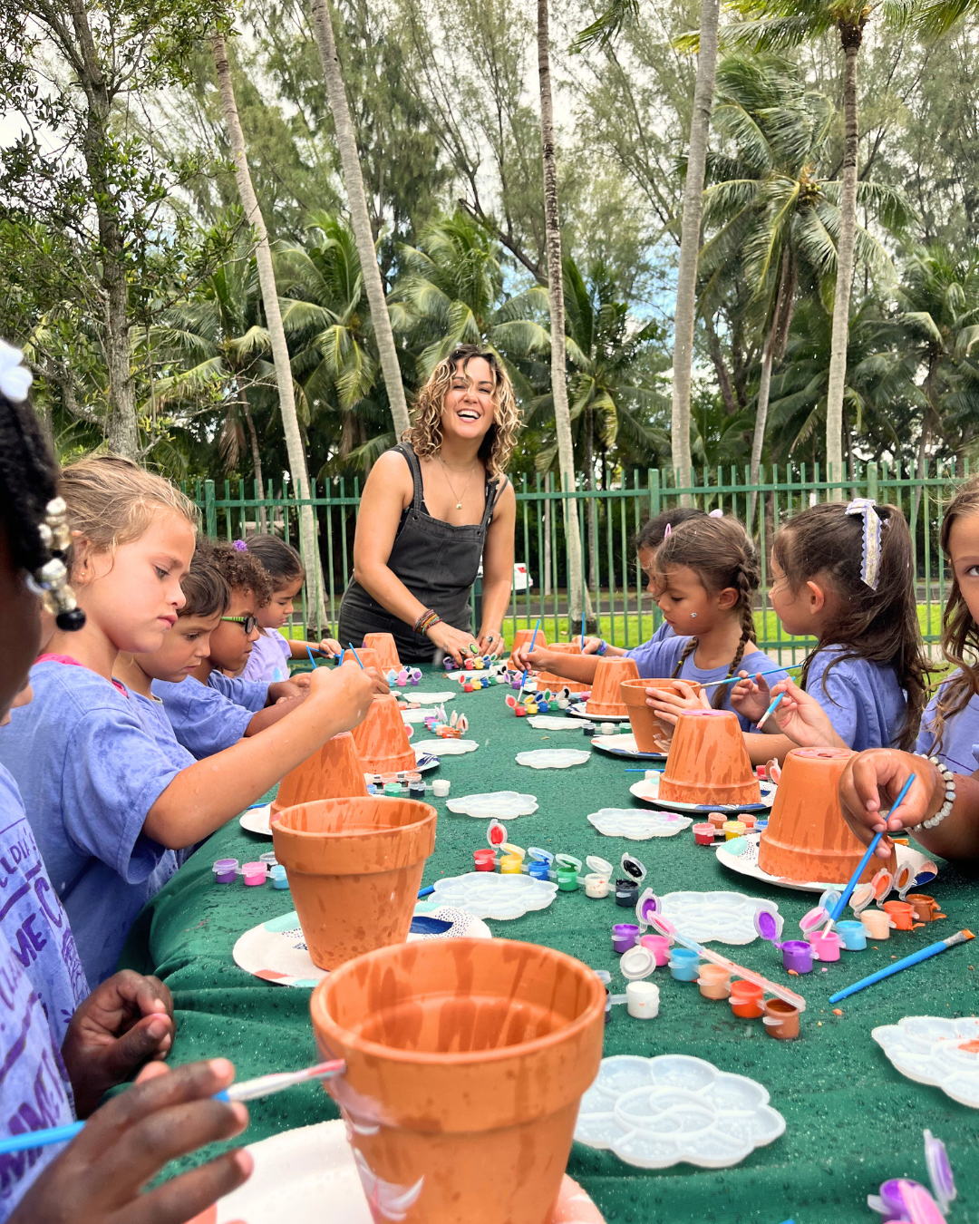 Children painting terracotta pots at a long outdoor craft table with a smiling instructor nearby