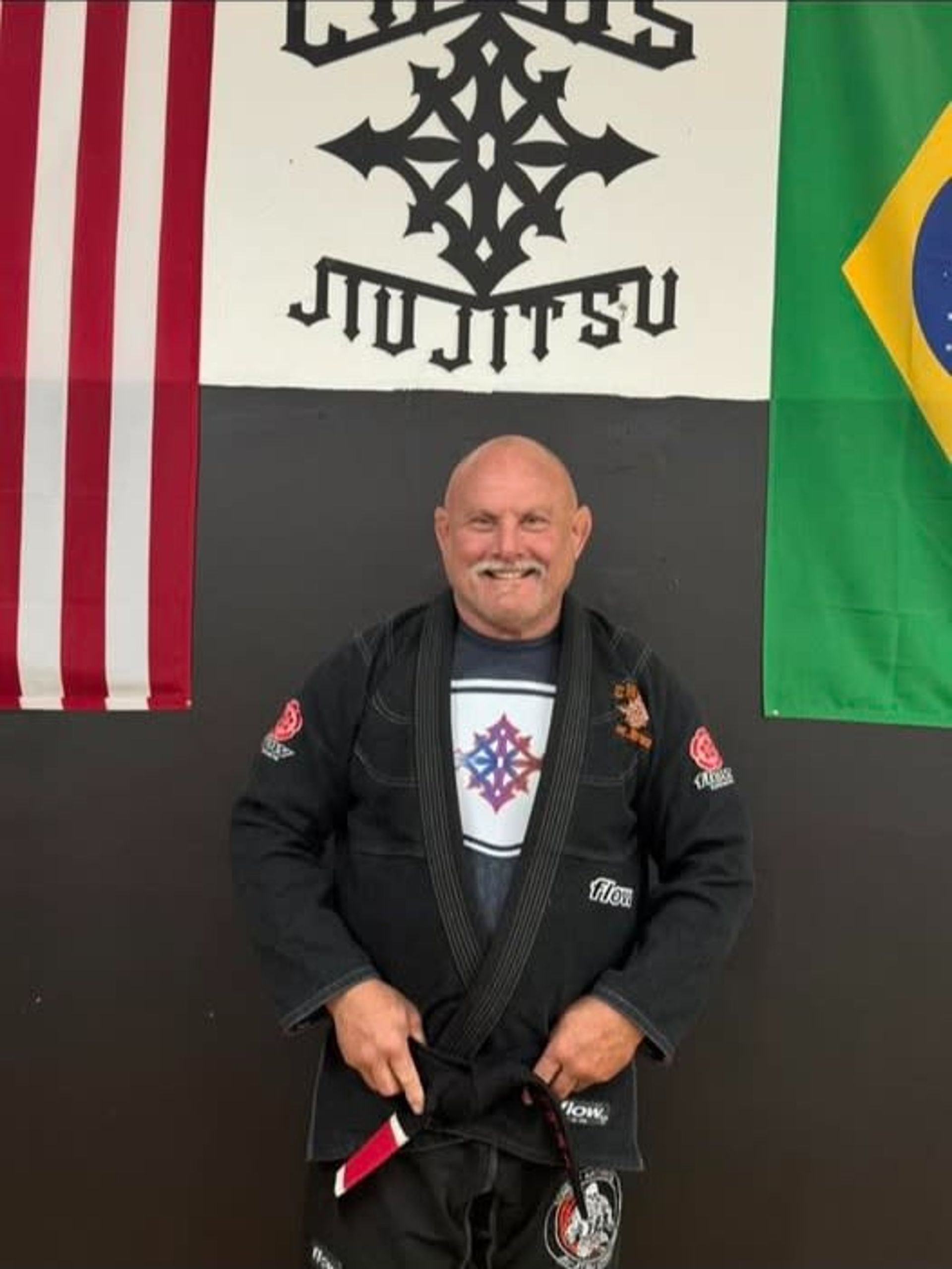 Man in a black Jiu-Jitsu gi stands in front of a banner and flags. He is smiling.