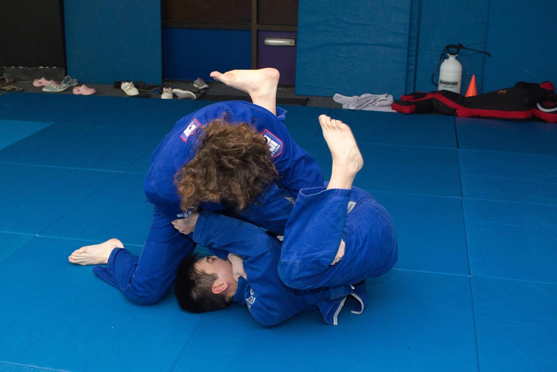 Two people in blue gis practicing jiu-jitsu on a blue mat.