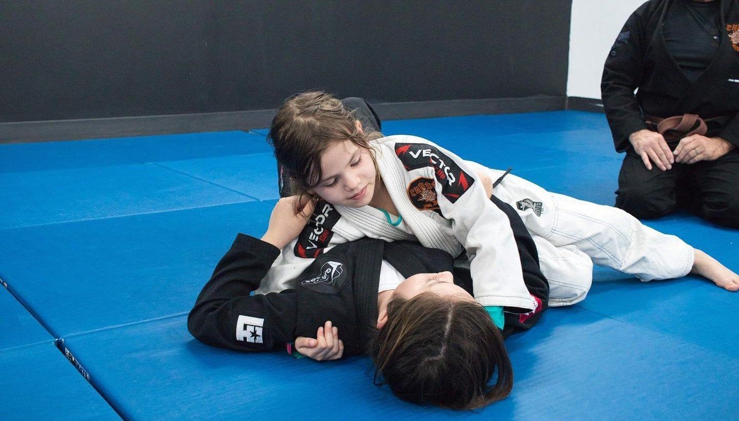 Two people in martial arts uniforms practice grappling on a blue mat.