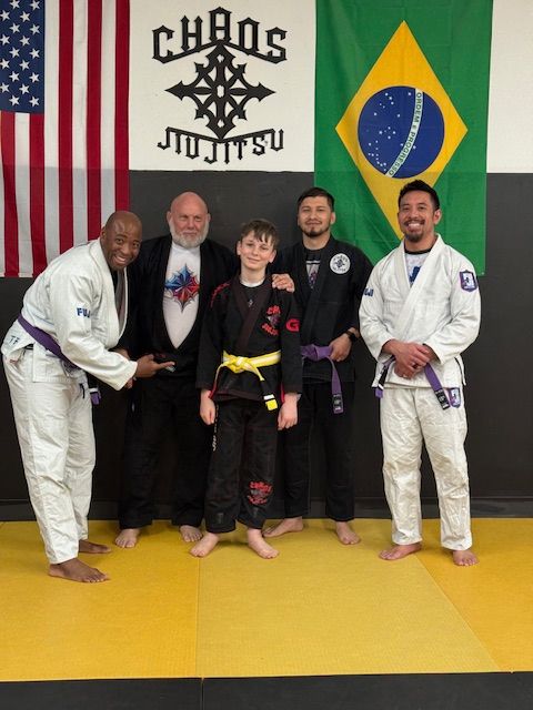 Five people in jiu-jitsu uniforms pose for a photo at a gym with American and Brazilian flags and a Chaos Jiu-Jitsu sign.