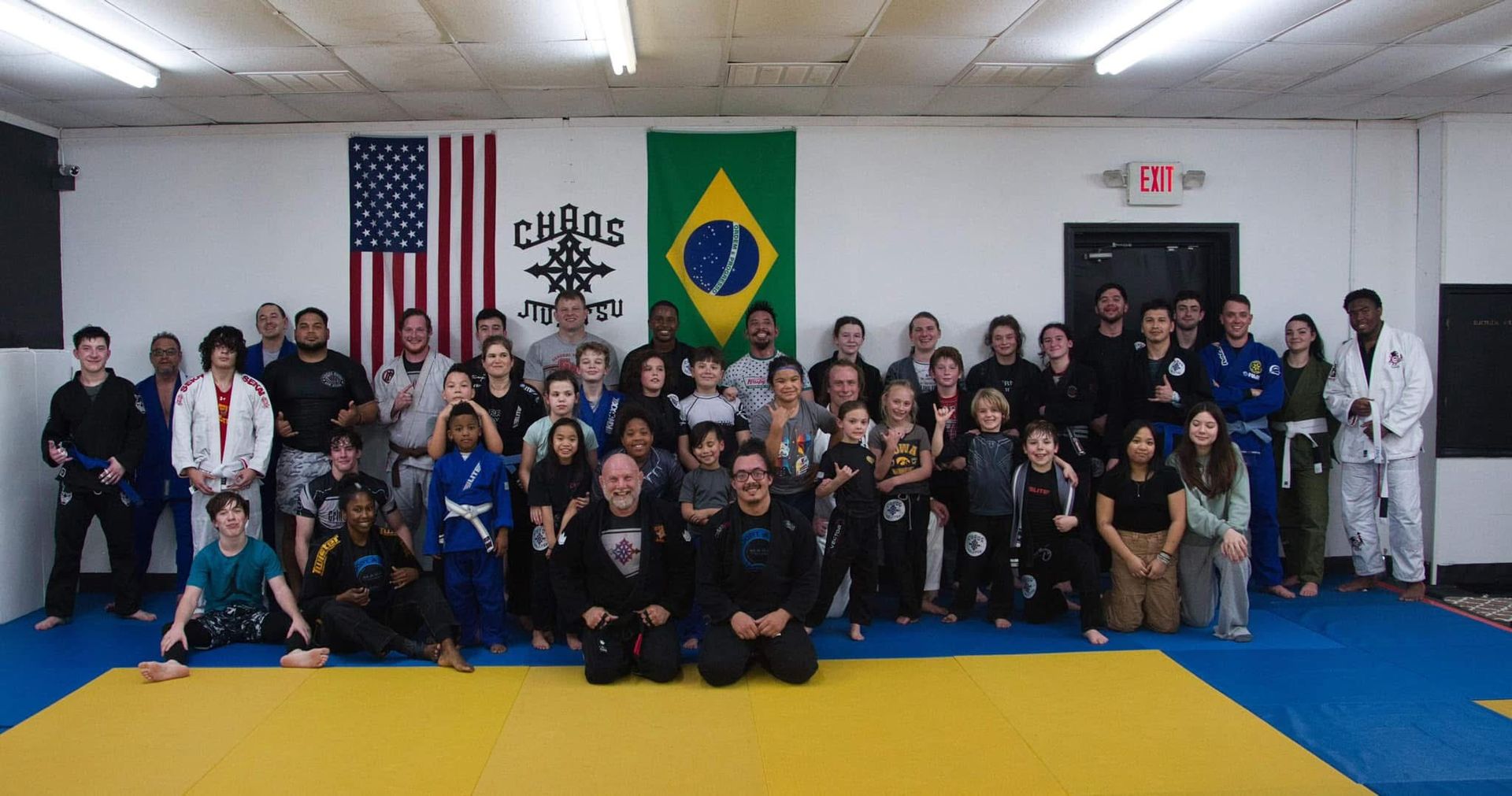 A diverse group of martial arts students posing for a photo in a gym in front of American and Brazilian flags.