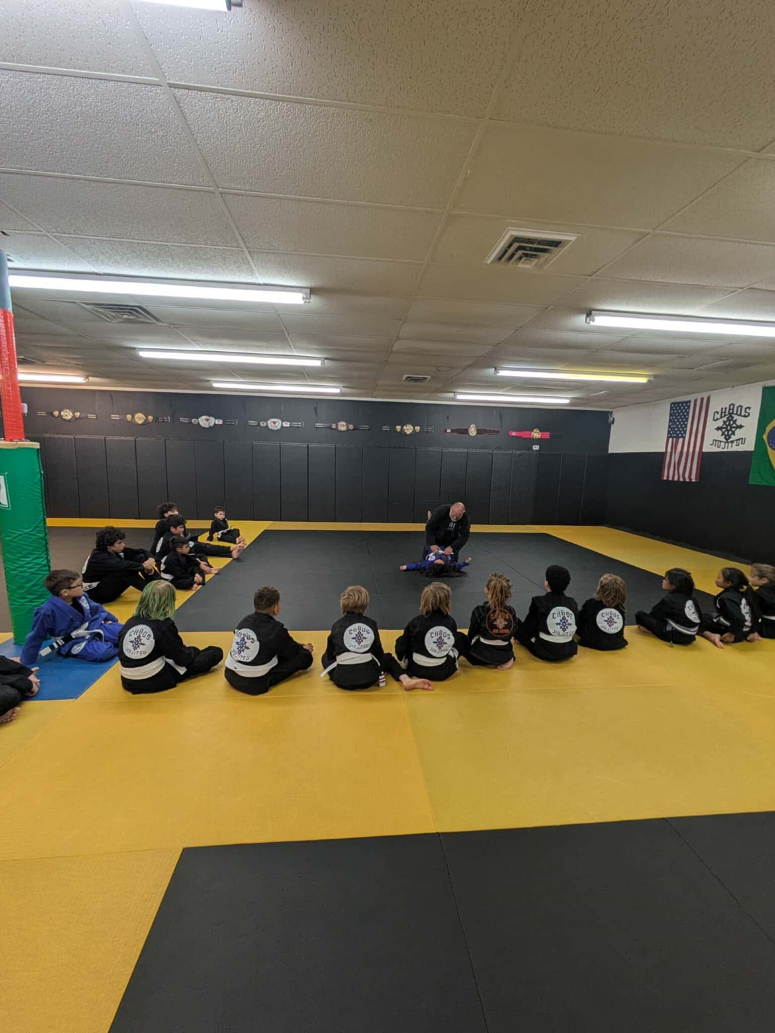 Students in martial arts uniforms sit in a semi-circle on yellow floor mats while an instructor demonstrates on the mat.