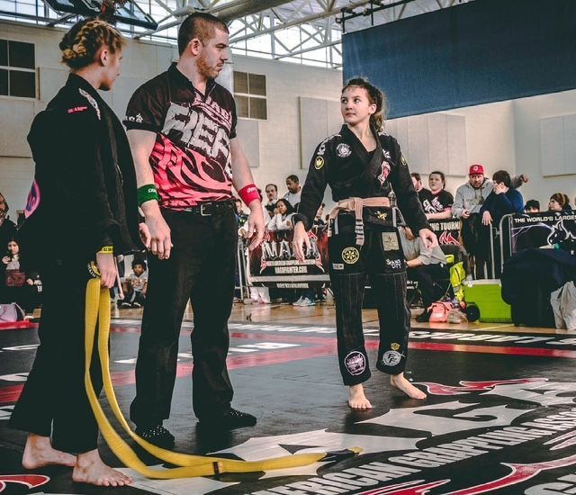 A referee stands between two competitors on a Brazilian Jiu-Jitsu mat in an indoor gym setting.