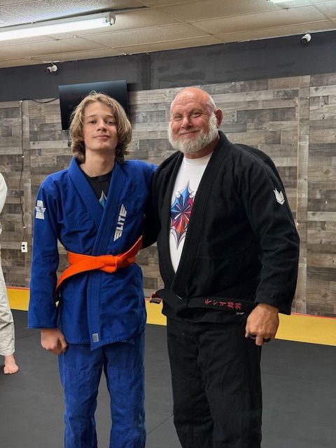 A student in a blue gi with an orange belt stands next to an instructor in a black gi in a martial arts training gym.