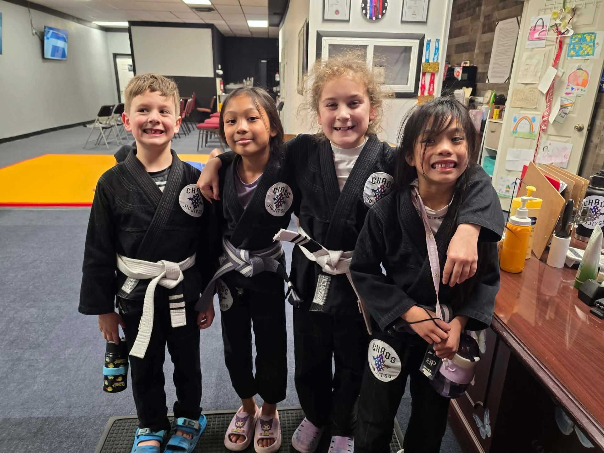 Four children in black martial arts uniforms and white belts smiling together in a dojo setting.