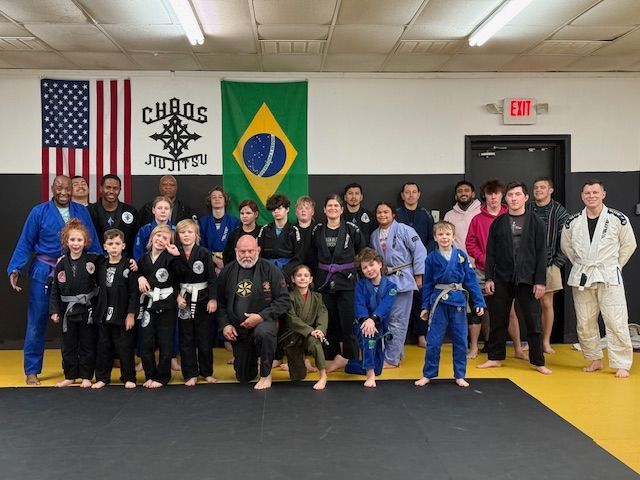 A group of martial artists in uniforms pose on a yellow mat in front of American and Brazilian flags in a training gym.