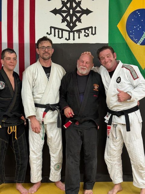 Four individuals in Jiu-Jitsu gis stand in front of flags at a gym; one person gives a thumbs-up.