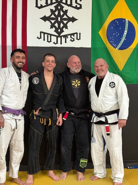 Four martial artists in Jiu-Jitsu gis stand smiling in front of a Chaos Jiu-Jitsu banner, a US flag, and a Brazil flag.