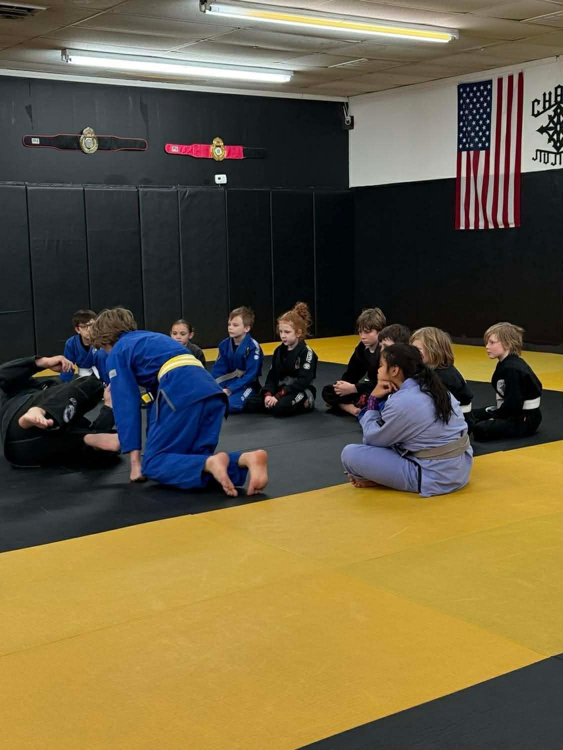 Children in martial arts uniforms sit and practice grappling on a padded floor inside a gym with an American flag.
