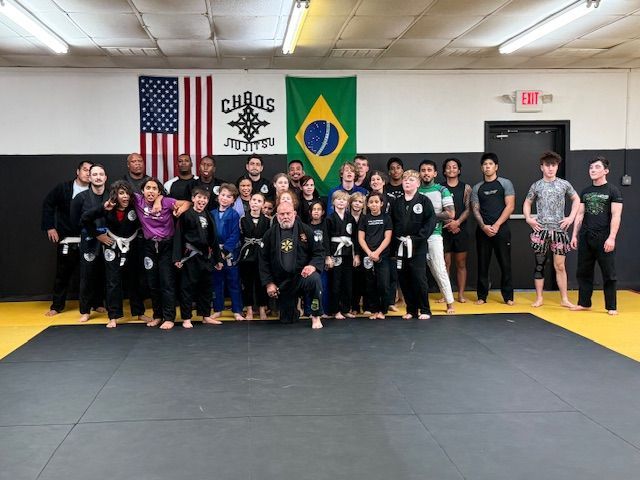 A group of people in martial arts uniforms posing for a photo in a gym with American and Brazilian flags on the wall.