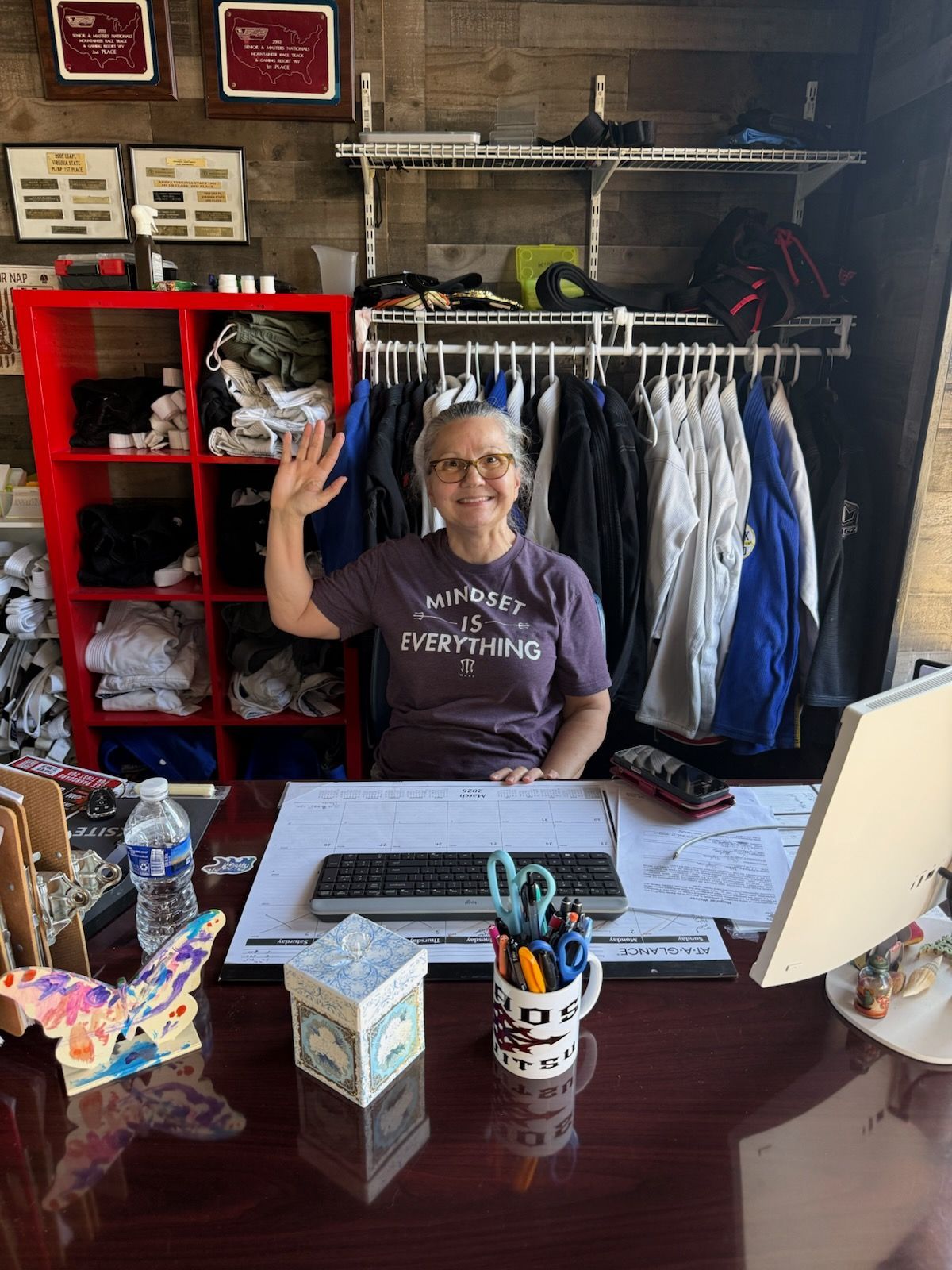 Woman waving from behind a desk in a shop; red shelves, clothing rack, and colorful items.
