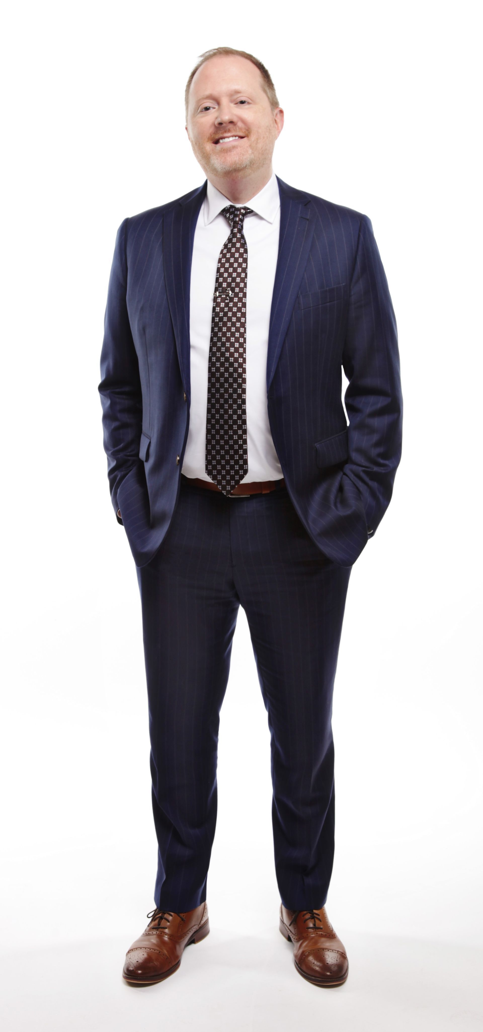 Man in a dark suit with hands in his pockets, smiling. White background.