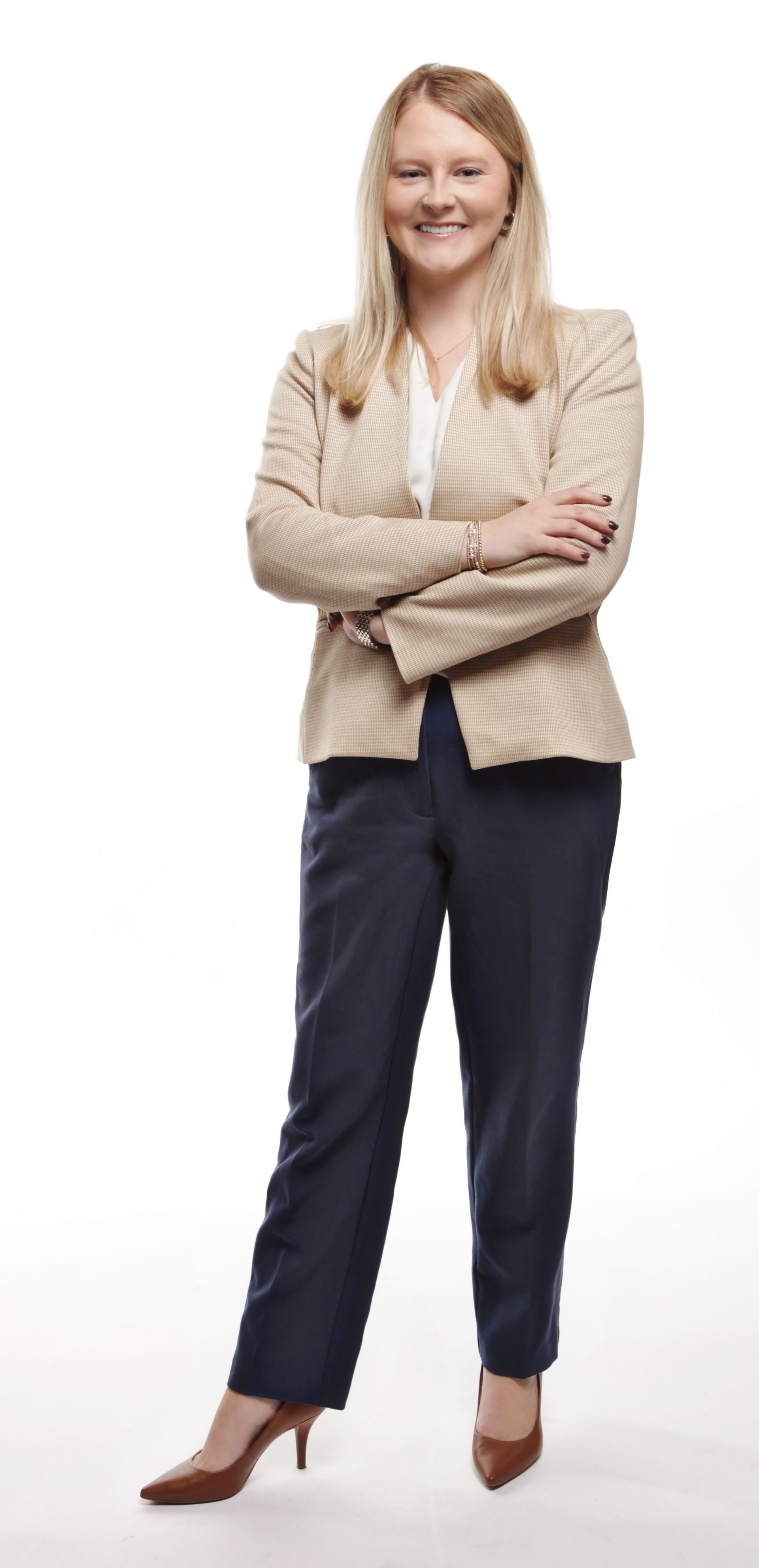 Woman wearing a patterned jacket, white shirt, and dark pants. Her arms are crossed, she smiles, and is wearing heels.