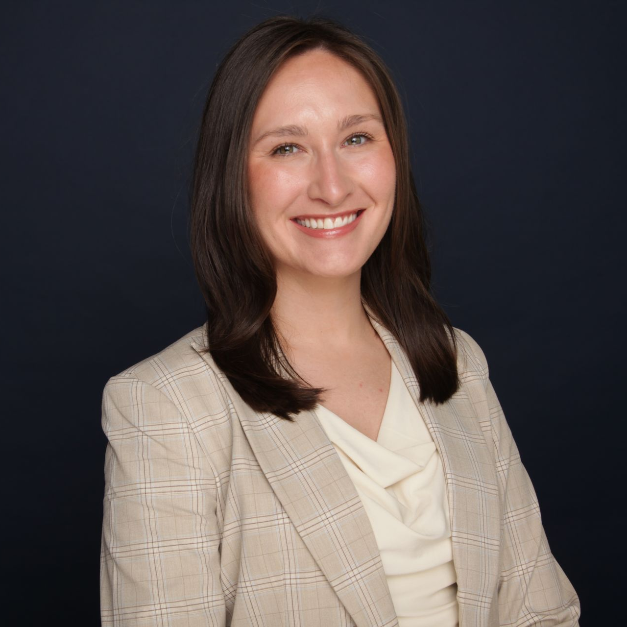 Woman with long brown hair smiling, wearing a black blazer and white shirt, against a dark blue background.