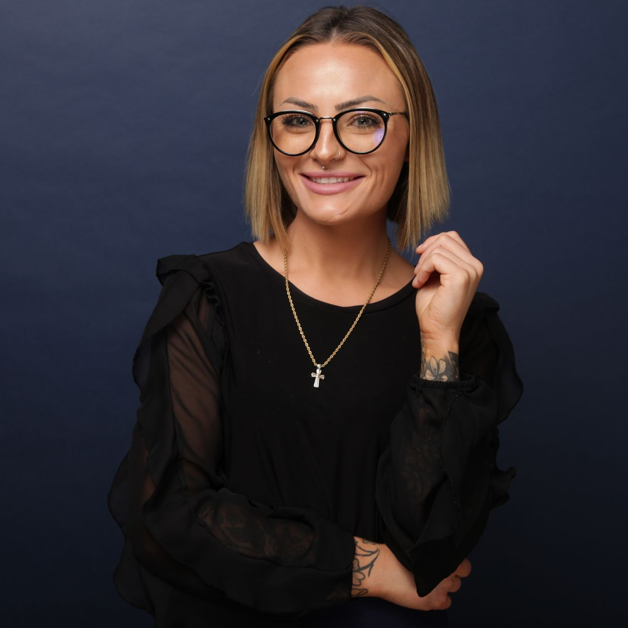 Woman with long brown hair, smiling, wearing a dark blue blouse against a dark background.