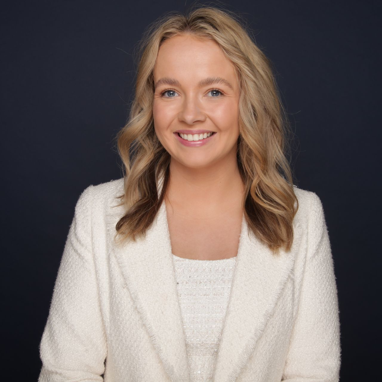 Woman with blonde hair wearing a beige blazer smiles in a studio setting.