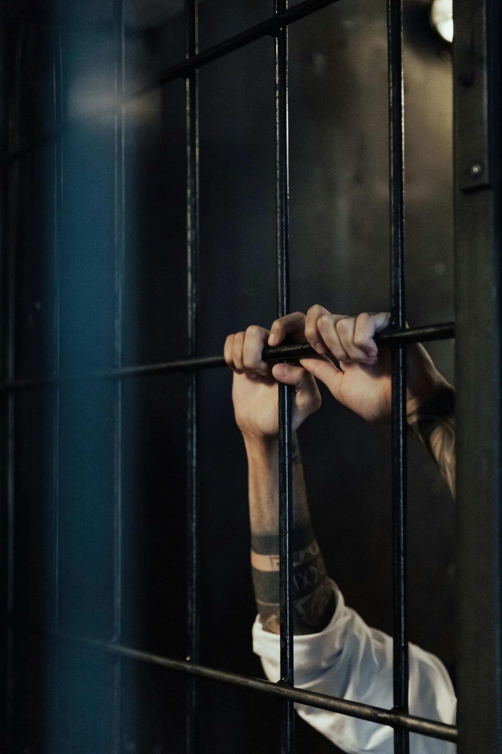 Hands holding jail bars. Dark setting, arm with tattoos, white shirt.
