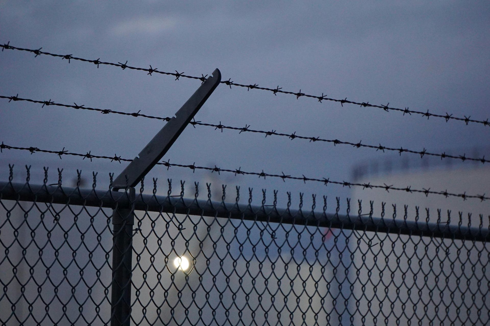 Chain-link fence with barbed wire on top against a twilight sky; a building is visible in the background.