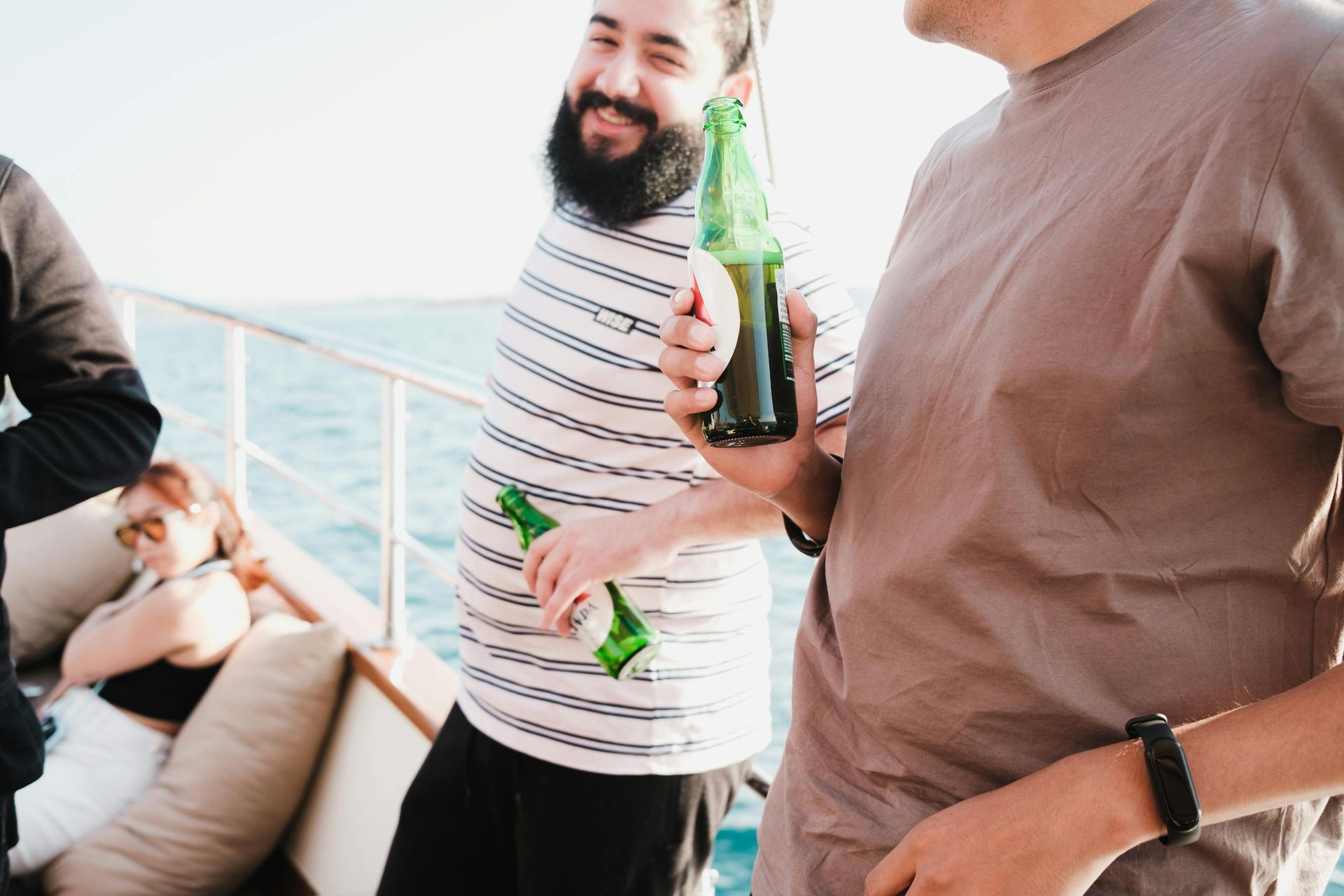 Man with beard holding beer bottle on boat, smiling. Another man nearby, sunlit sea in background.