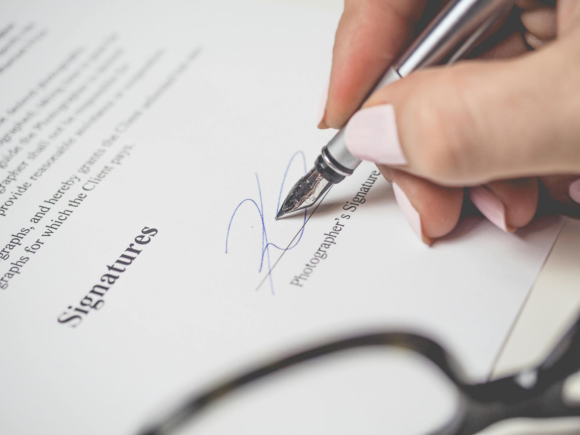 A hand with pink nails signs a document with a fountain pen, next to eyeglasses.