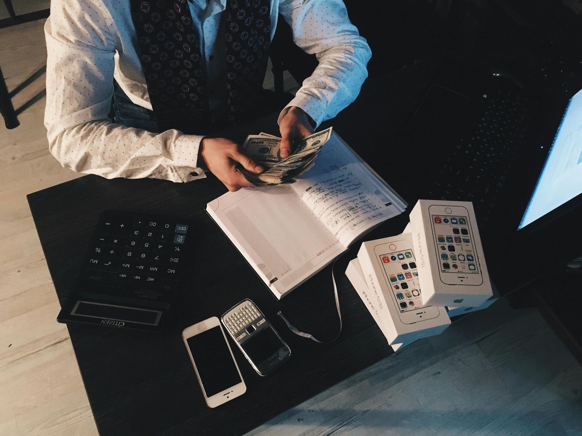 Person counting cash at a desk with notebook, calculator, phones, and stacked papers.