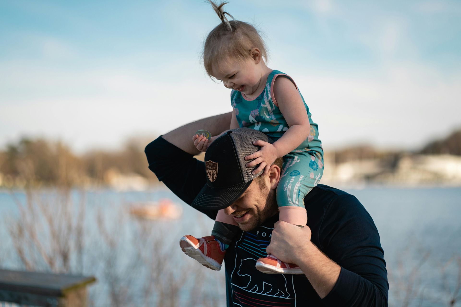Man carrying a child on his shoulders near a lake. The child is laughing, looking down. Clear blue sky.