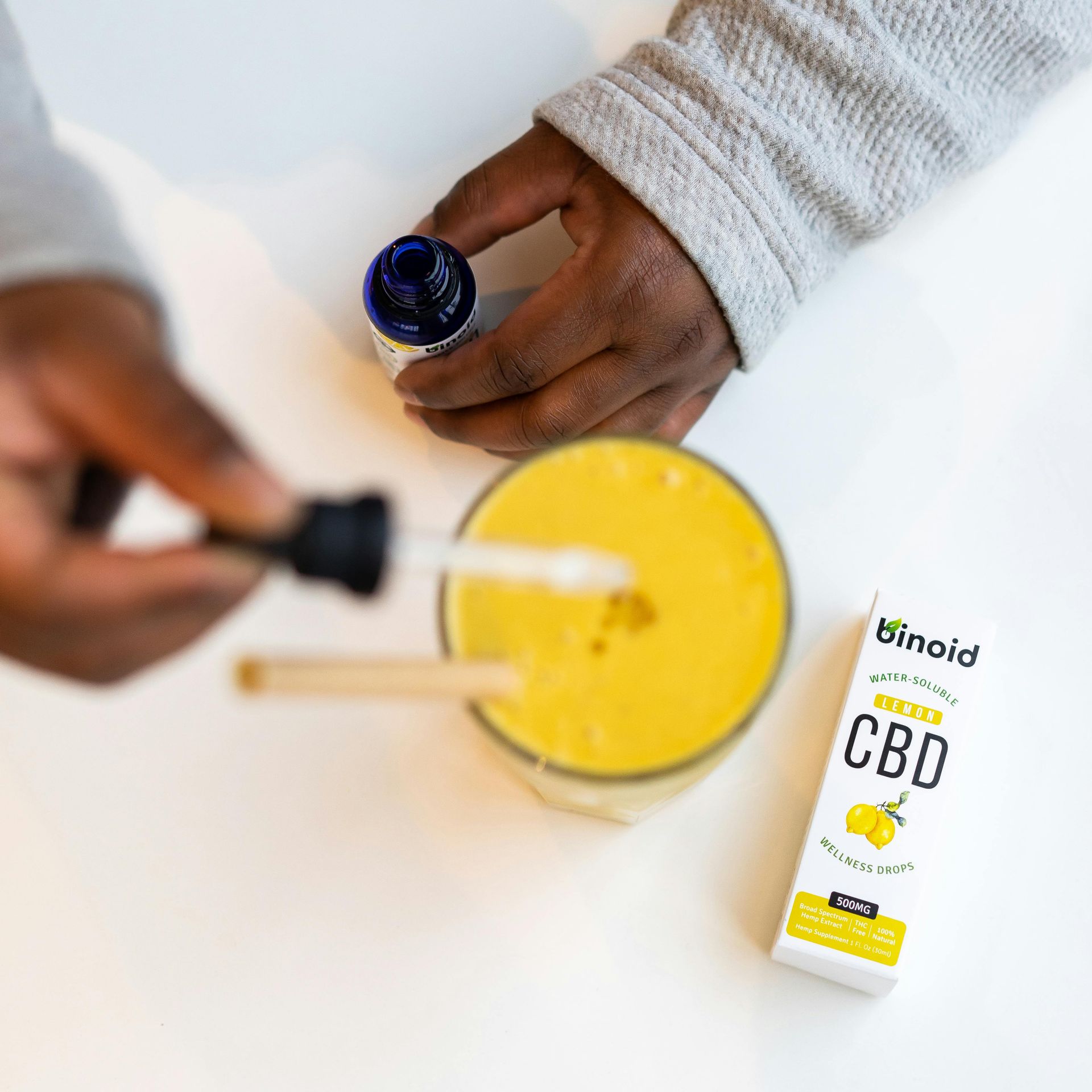 Person adding CBD drops to a yellow drink. White table, dropper, and a CBD oil box in view.