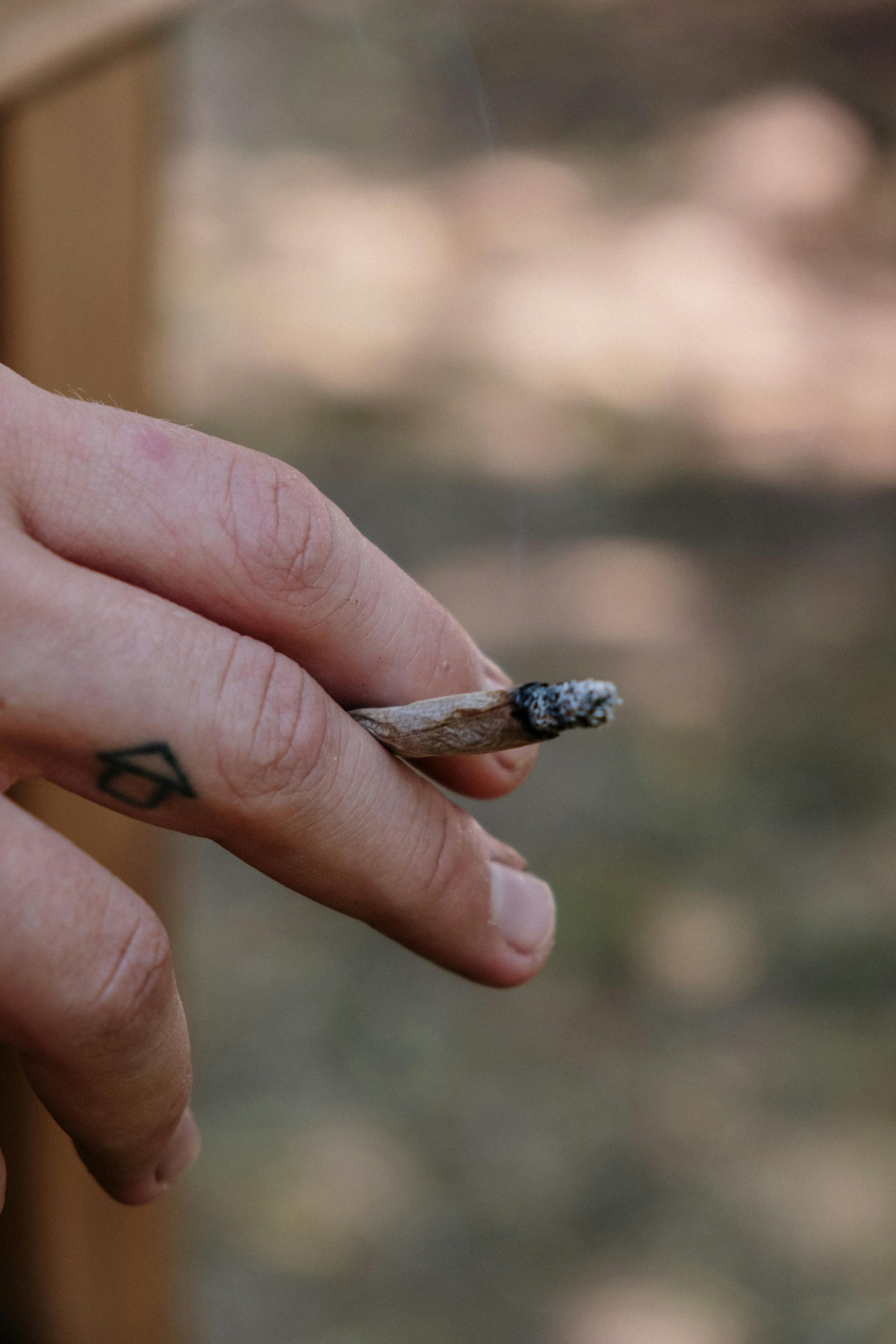 Hand holding a lit, rolled cigarette. A small tattoo is visible on the hand. Blurred background.