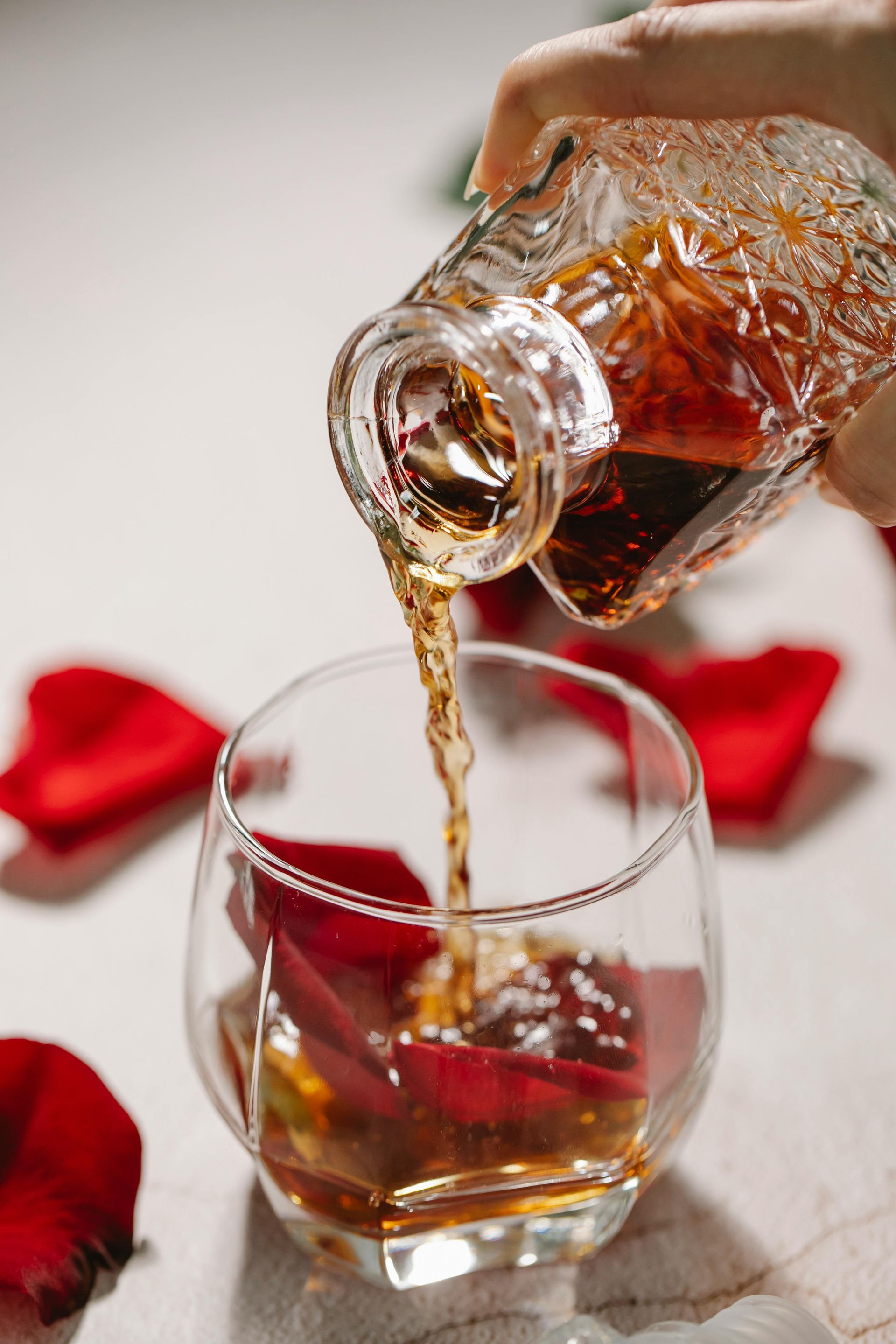 Whiskey pouring from a patterned bottle into a glass with red rose petals; white tablecloth.