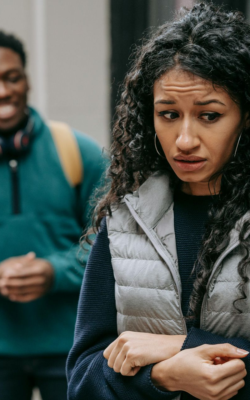 Woman with concerned expression, looking down. Man with headphones, appears to be smiling in the background. Outdoor setting.