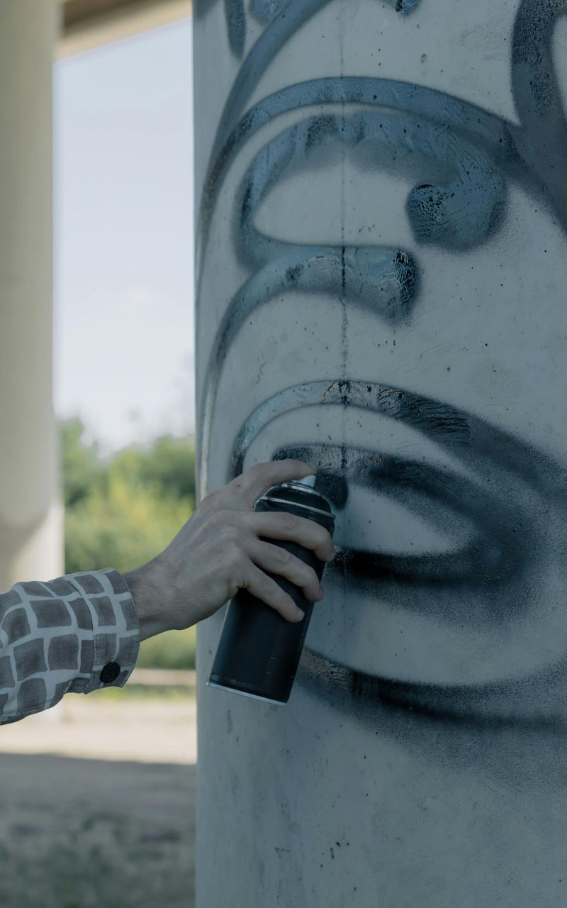 Person spray-painting graffiti on a concrete pillar outdoors.