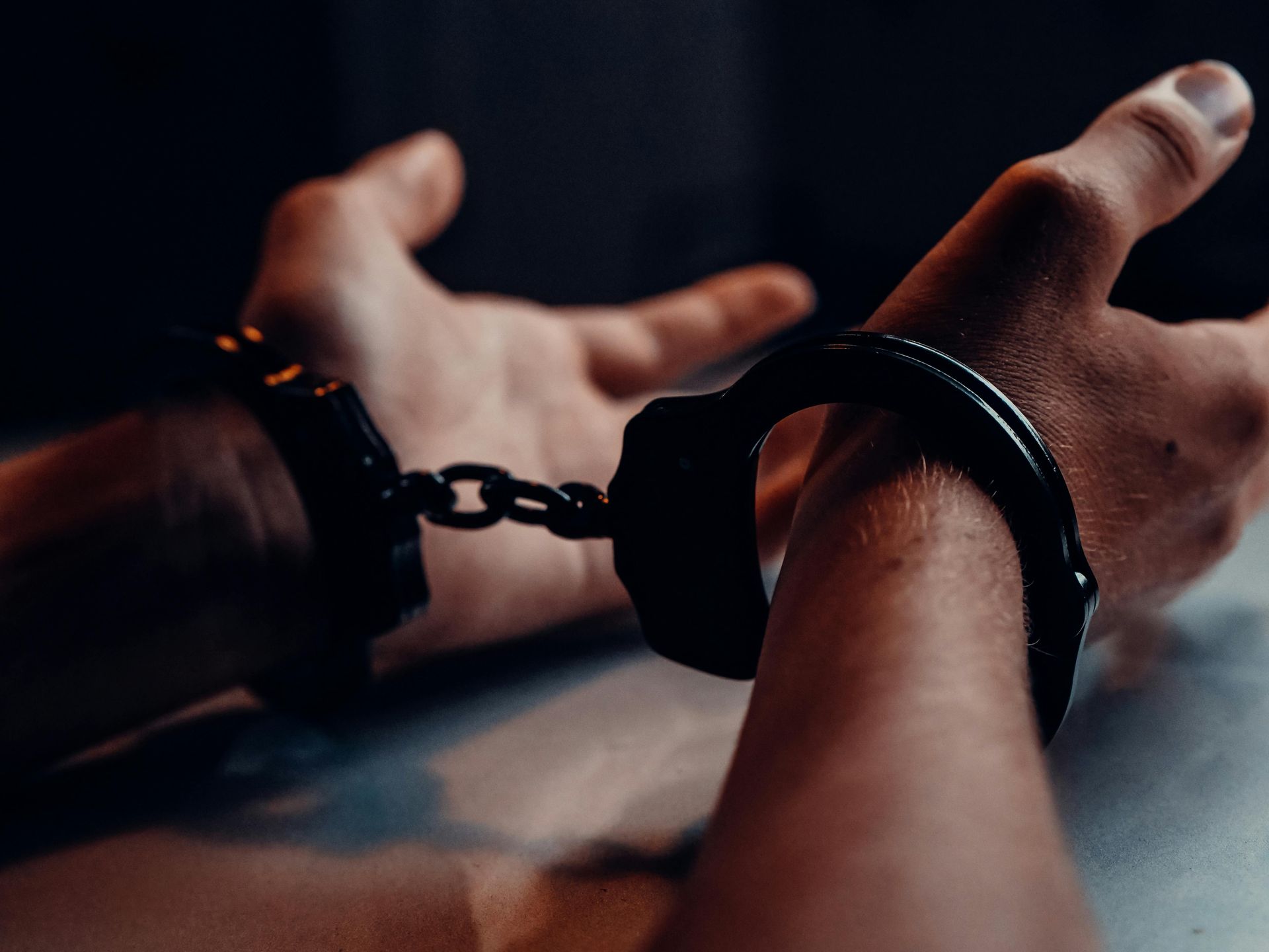 Hands in handcuffs, wrists chained, dark background.