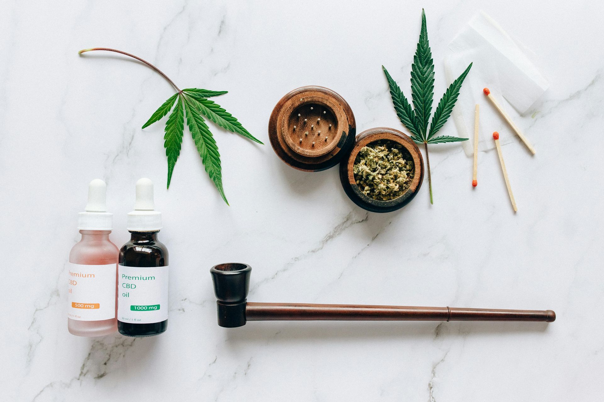 Marijuana setup: leaves, grinder with bud, pipe, dropper bottles, matches, and rolling papers on a white surface.