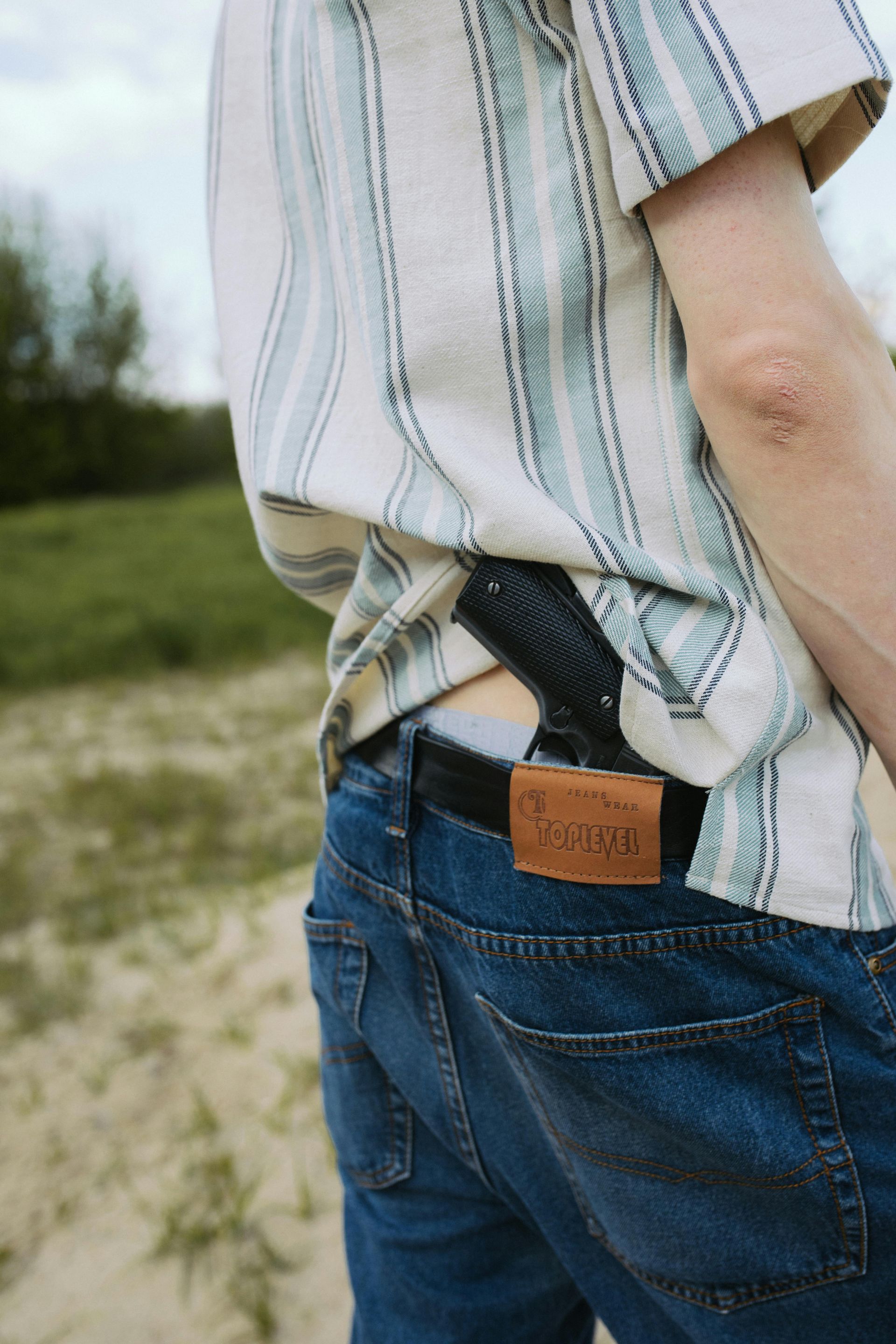 Person wearing jeans and a button-down shirt, a holstered handgun visible on their back. Outdoors on a dirt path.