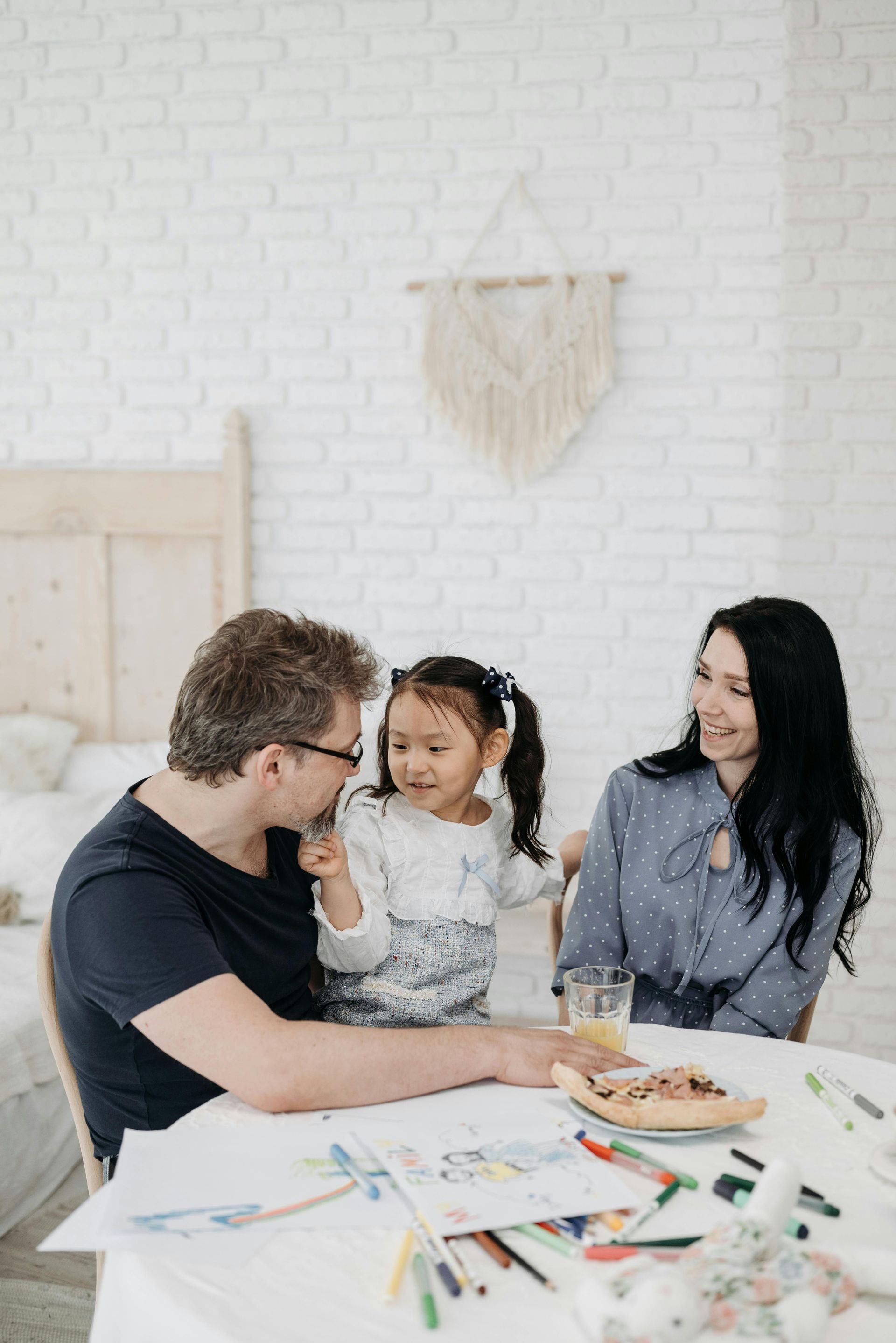 Family sitting at table, looking at each other, child with pigtails smiling. White brick wall background, art supplies.