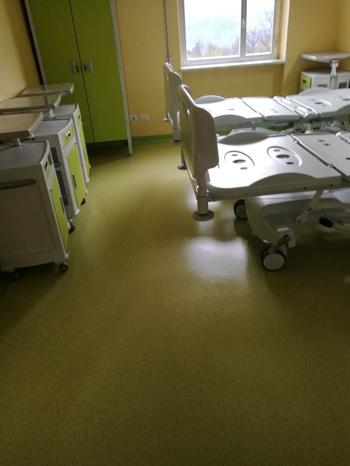 Stanza d'ospedale con due letti, armadi e una finestra. Colori verde e giallo.
